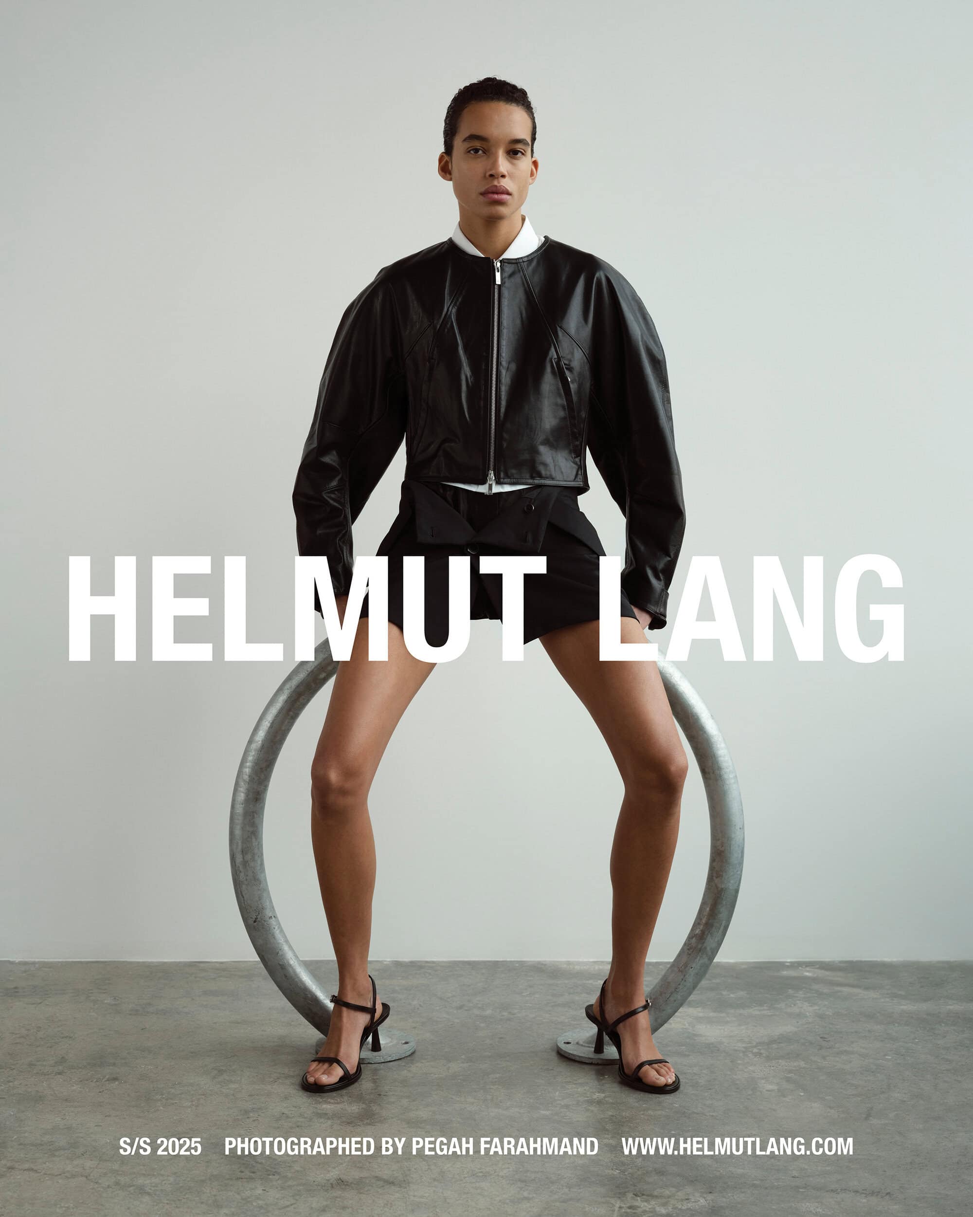 Model wearing a black leather jacket and shorts straddling a curved metal ring against a grey backdrop
