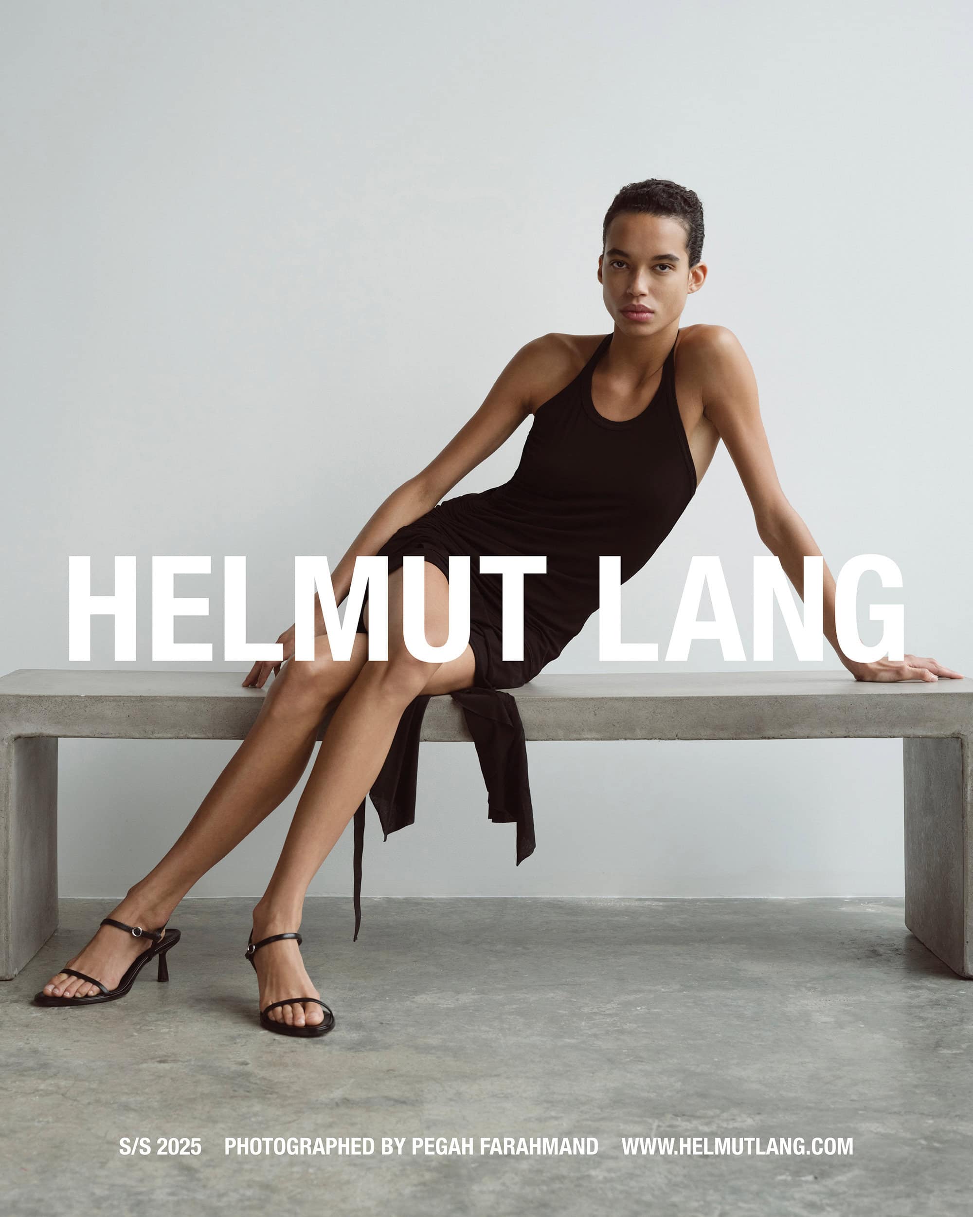 Model wearing a black halter dress and sandals seated on a concrete bench against a neutral wall