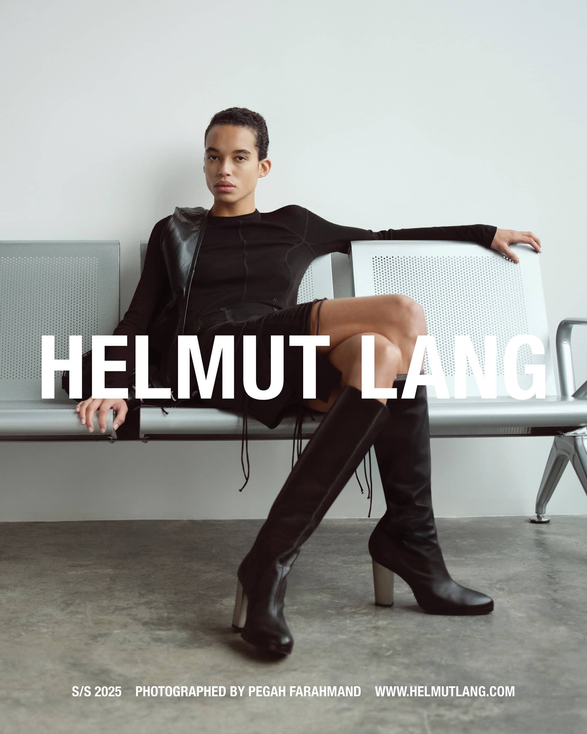 Model wearing black long-sleeve top and high-heeled boots seated on a metal bench against a plain wall.