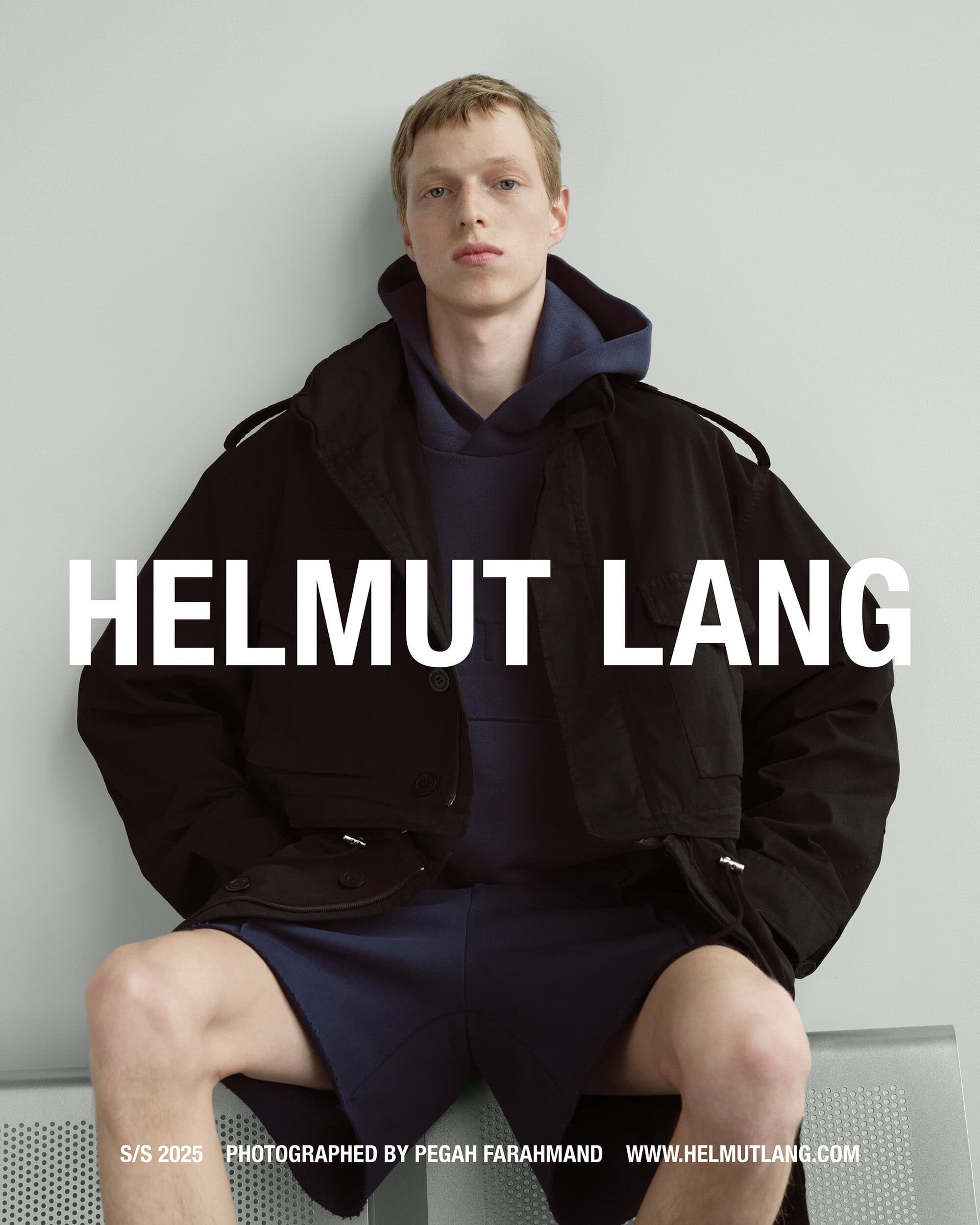 A male model seated wearing a dark jacket and blue hoodie against a light grey studio backdrop with Helmut Lang branding across the center.
