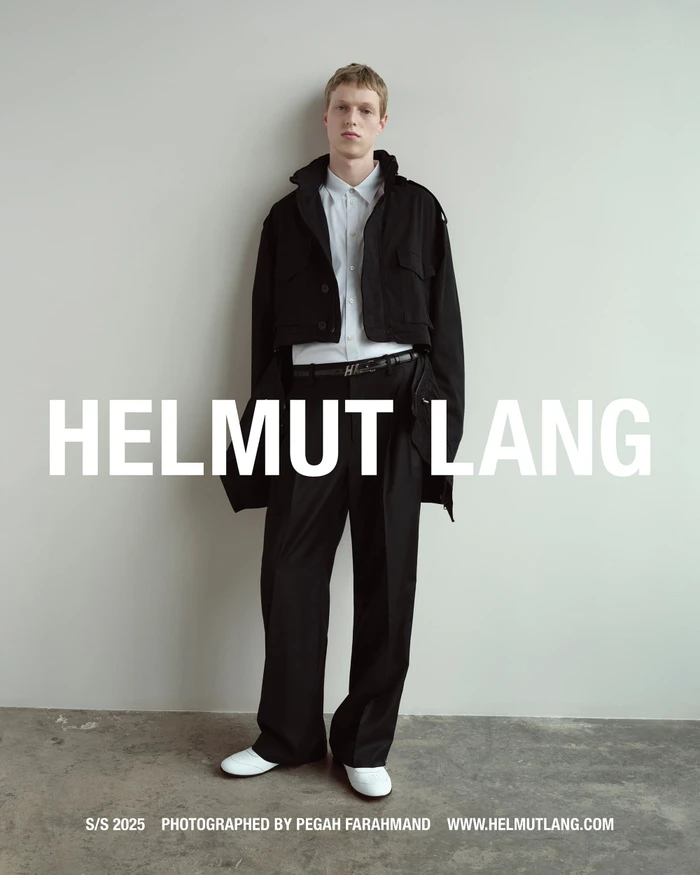 A young male model posing against a neutral wall in a black jacket, white shirt, black trousers, and white shoes.