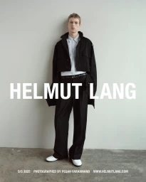 A young male model posing against a neutral wall in a black jacket, white shirt, black trousers, and white shoes.