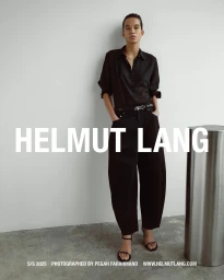 Model standing against a plain white wall wearing a black button-down shirt and black trousers with a metallic cylinder prop