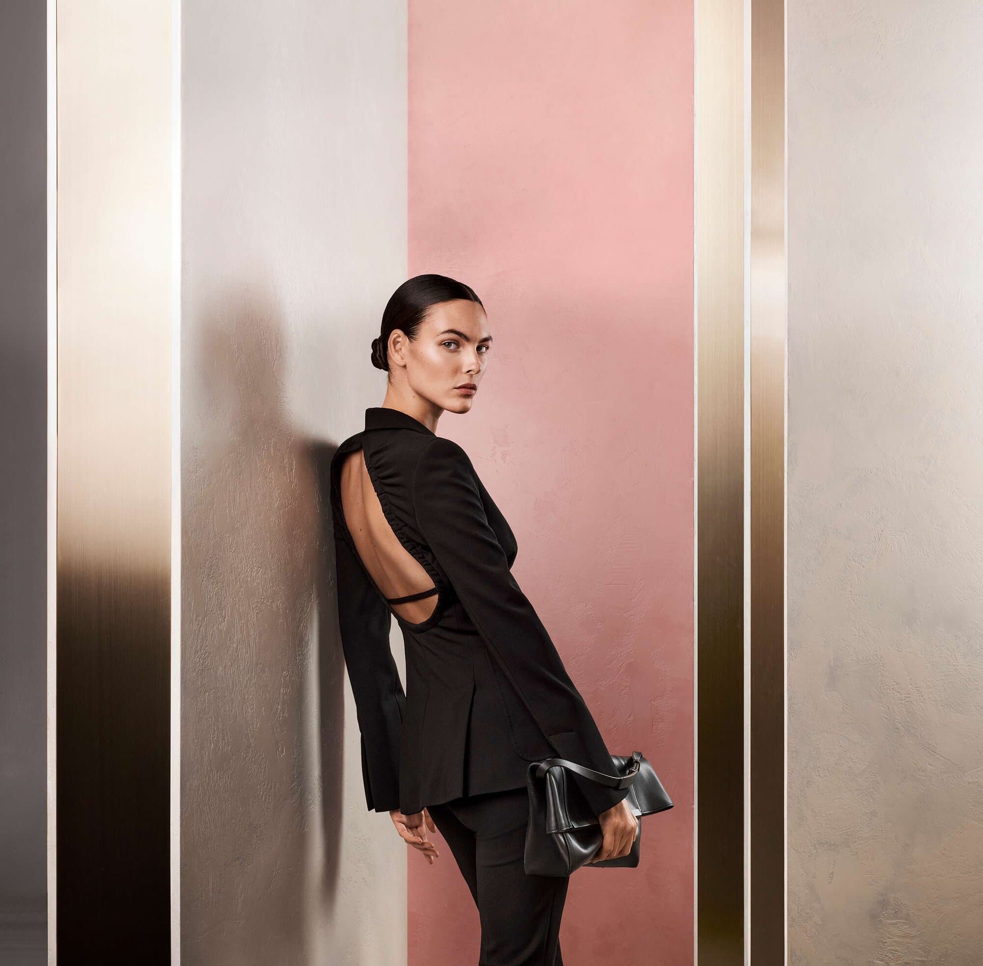 Model Vittoria Ceretti in a backless black blazer holding a handbag against a geometric wall in a Max Mara campaign.