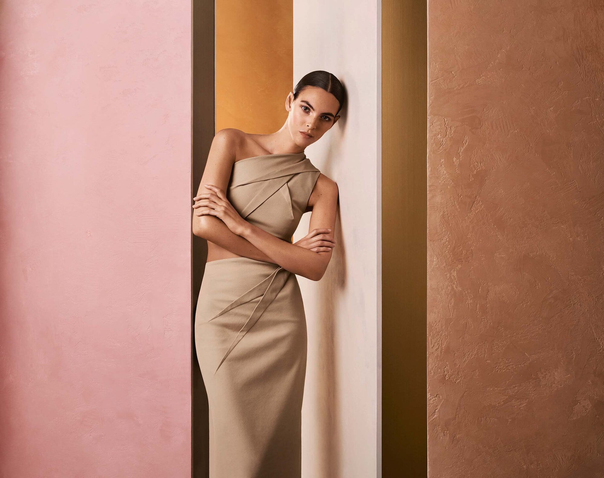 Model Vittoria Ceretti leaning against multi-toned architectural panels wearing a beige one-shoulder dress for Max Mara