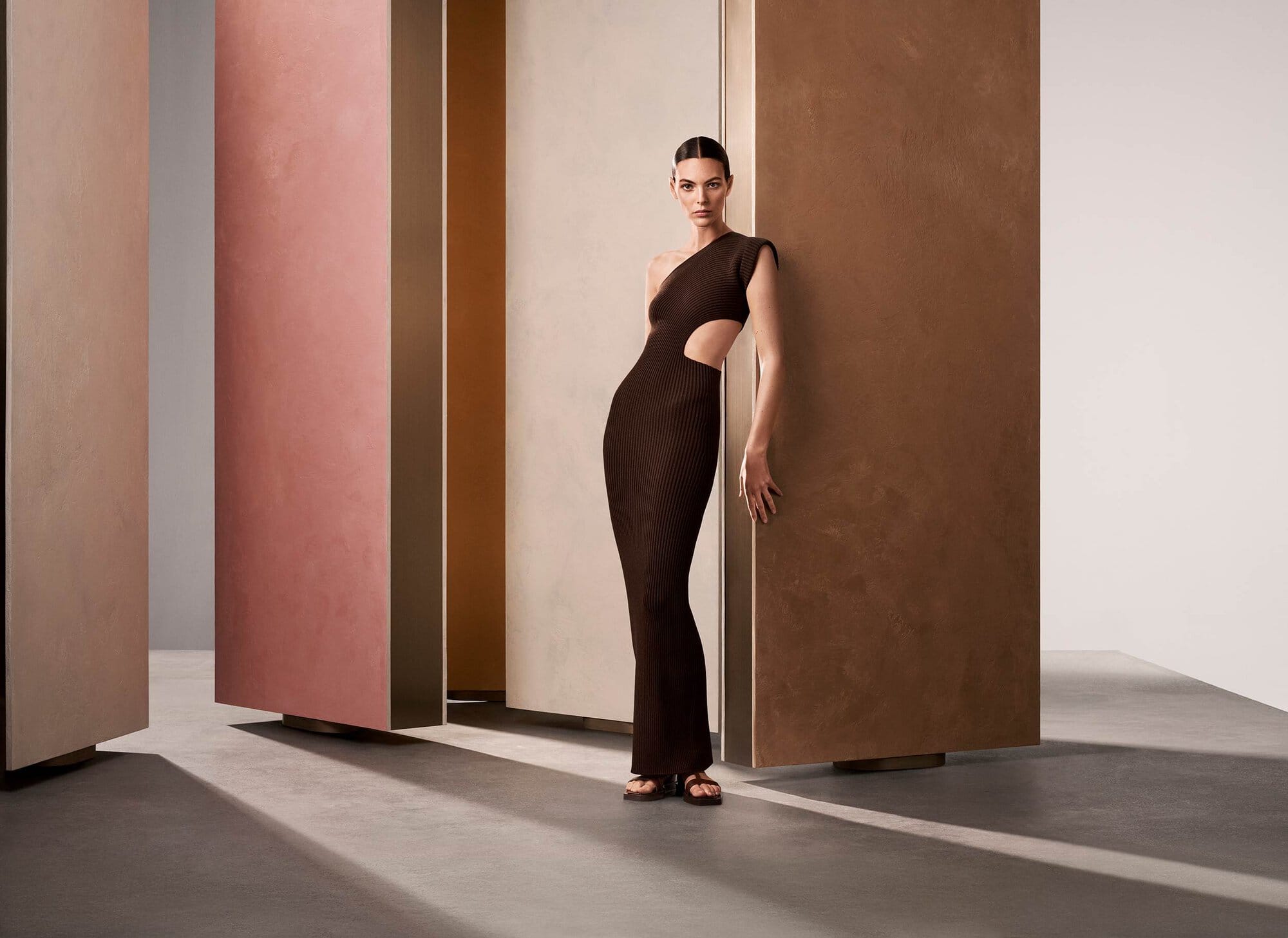 Model Vittoria Ceretti in a brown one-shoulder dress leaning against a tall textured panel in a minimalist architectural studio set.