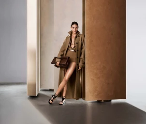 Model Vittoria Ceretti in a brown belted coat holding a clutch bag in a set-dressed studio environment.