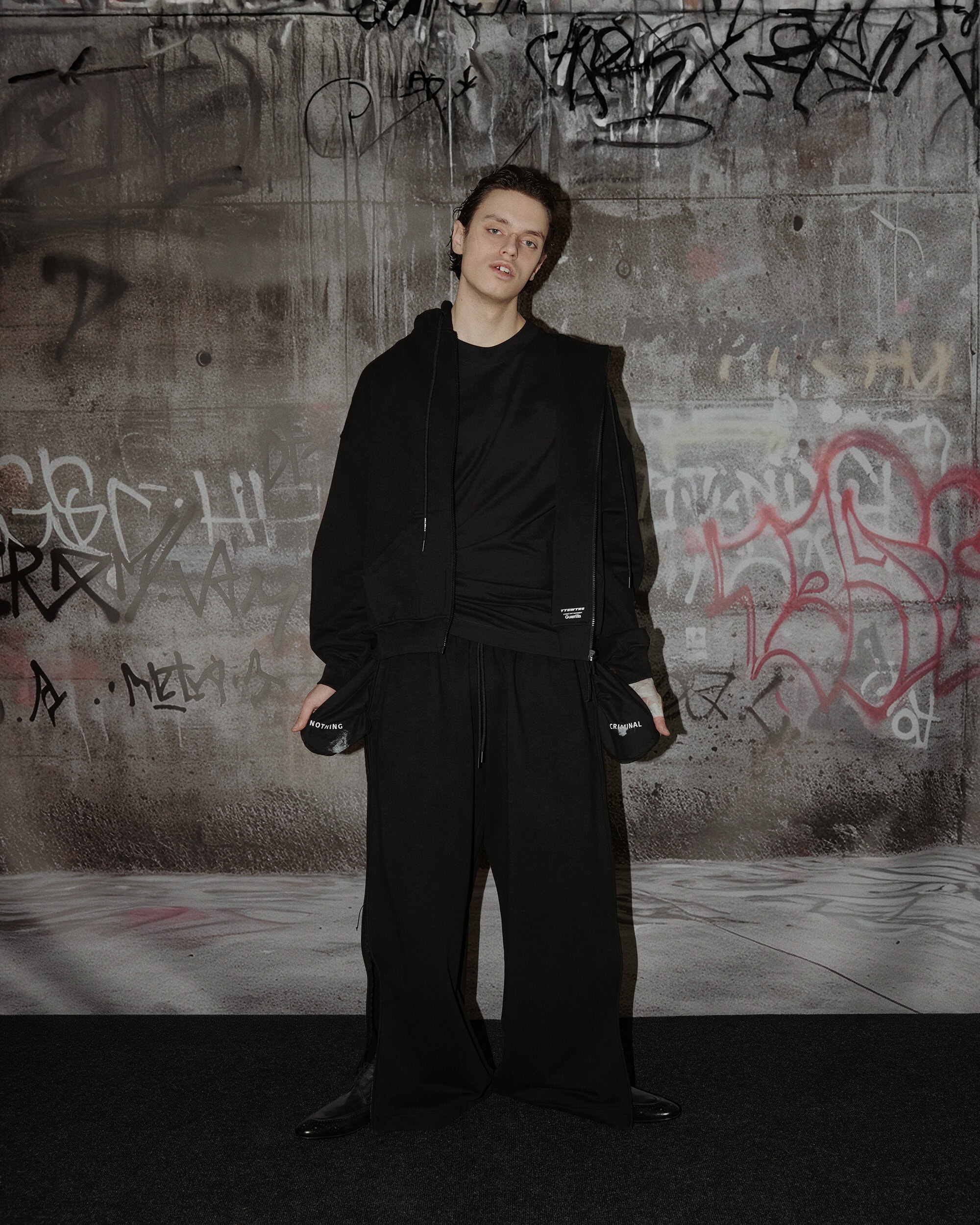 Young man in black hoodie and loose trousers standing against a graffiti-covered concrete wall for TTSWTRS Youth campaign