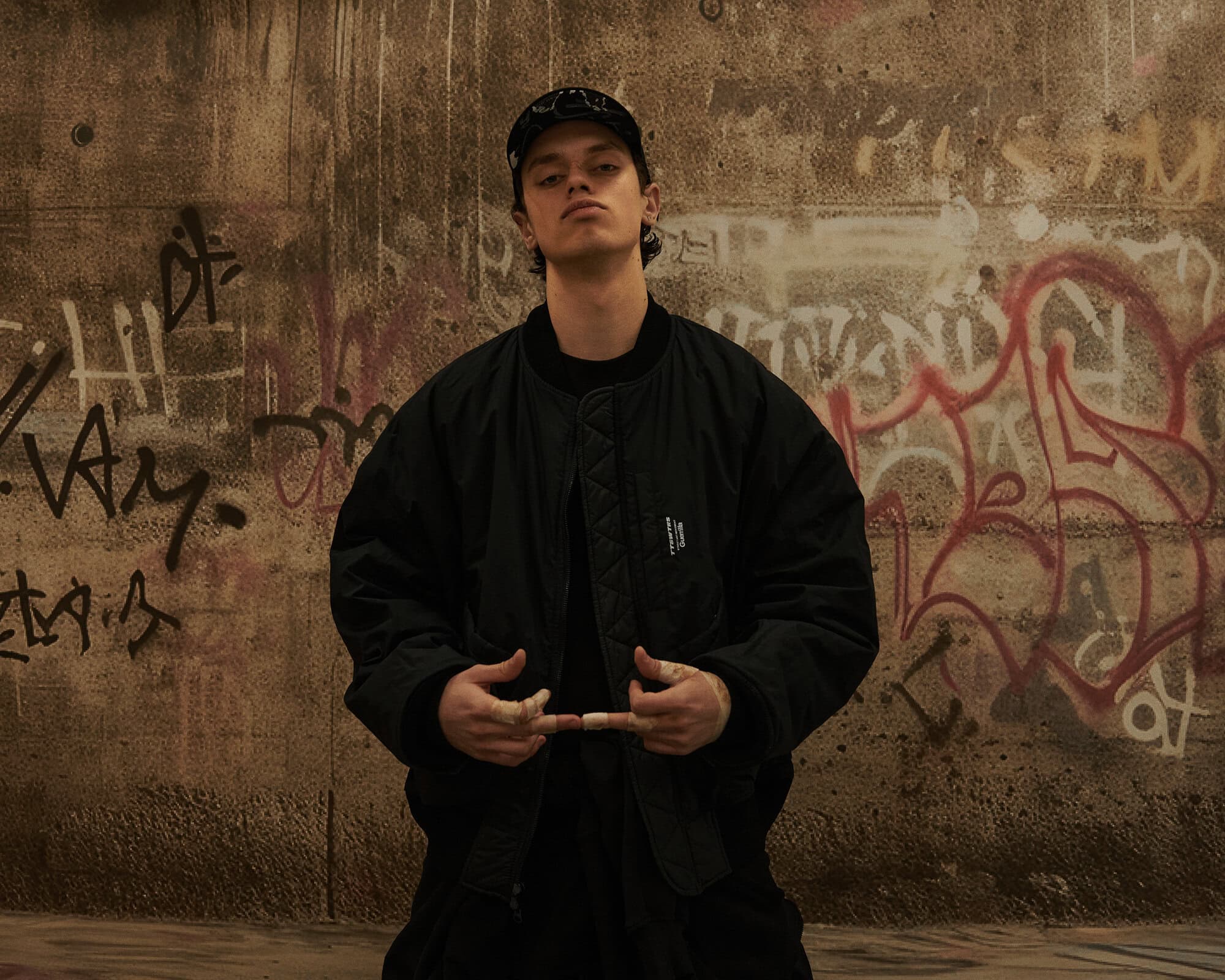 A young man wearing a black cap and jacket stands against a textured concrete wall with graffiti.