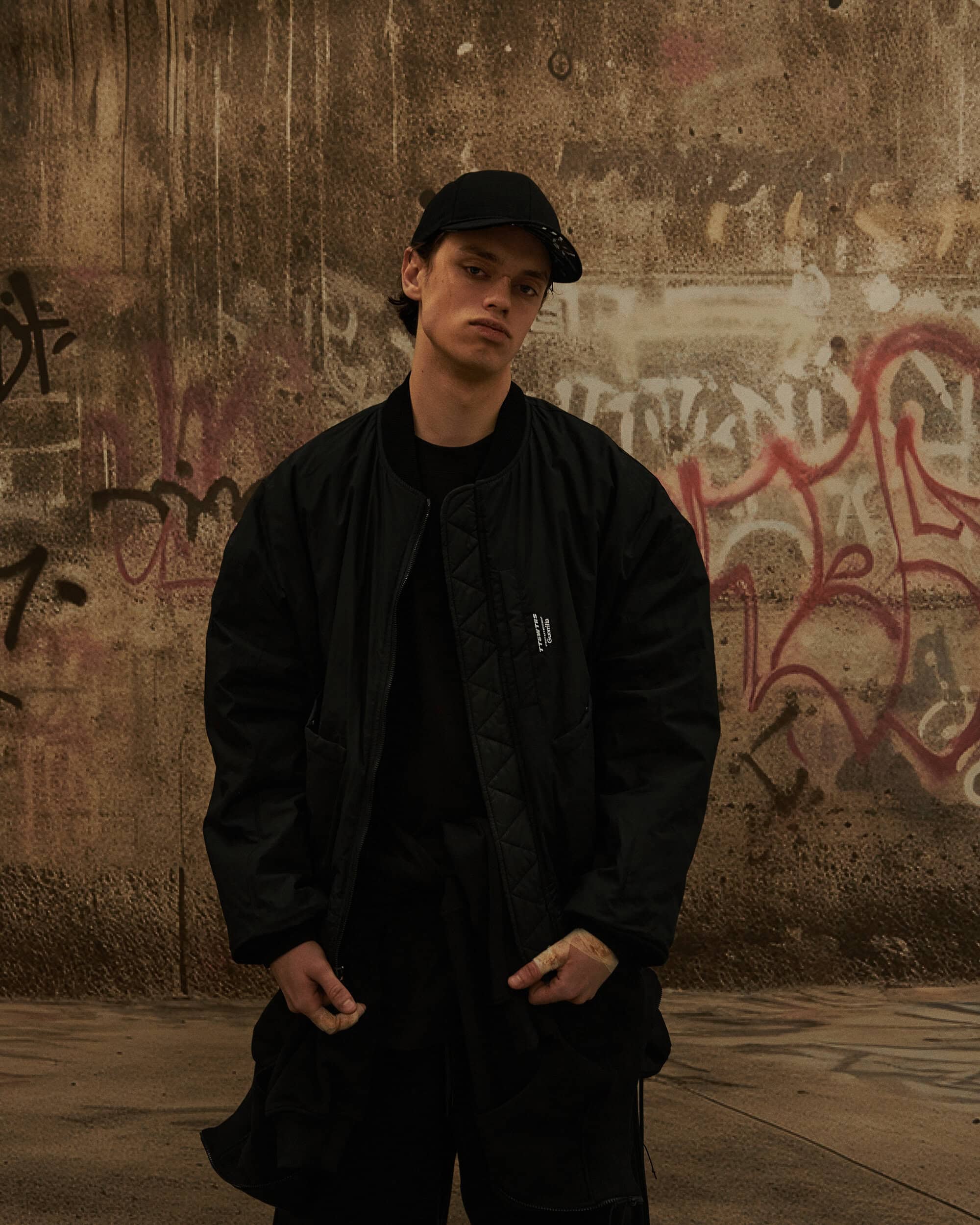 Young man wearing a black bomber jacket and baseball cap standing against a graffiti-covered concrete wall.