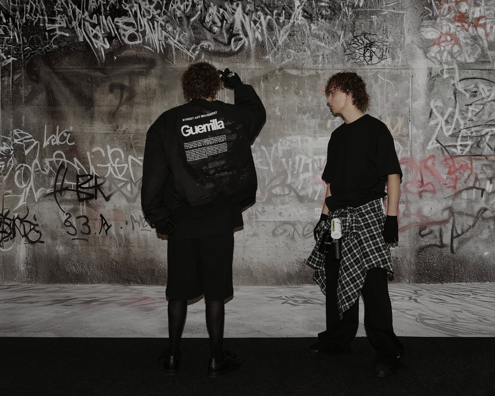 Two people in black streetwear standing in front of a graffiti-covered concrete wall for TTSWTRS Spring 2025 campaign.
