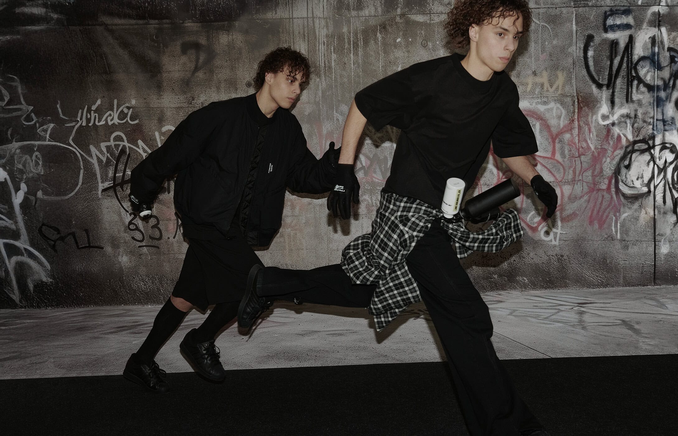 Two young male models in black outfits running against a gritty concrete wall with graffiti backdrop for TTSWTRS Youth campaign