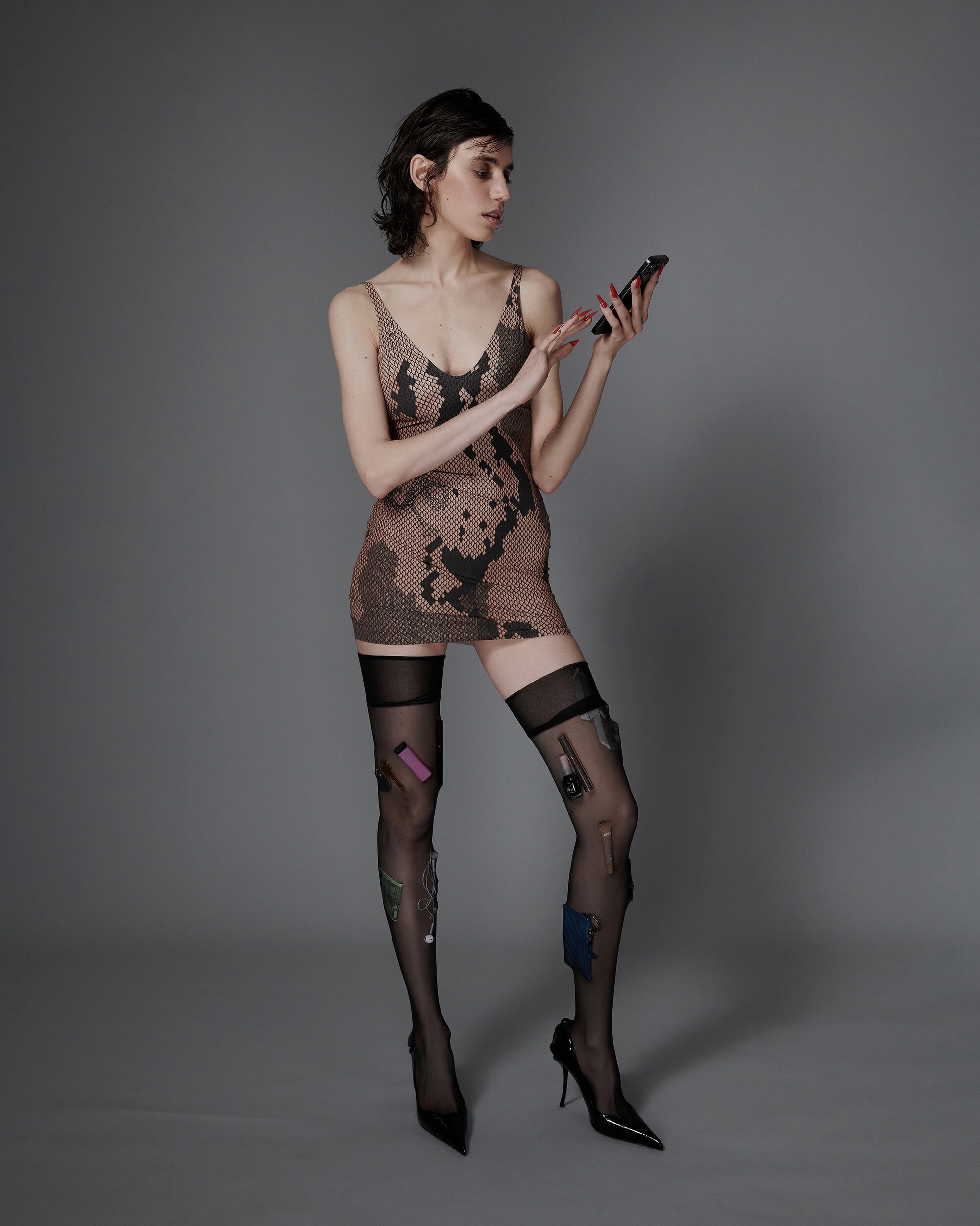 Model in snakeskin print mini dress and sheer stockings holding a smartphone against a grey studio seamless backdrop.