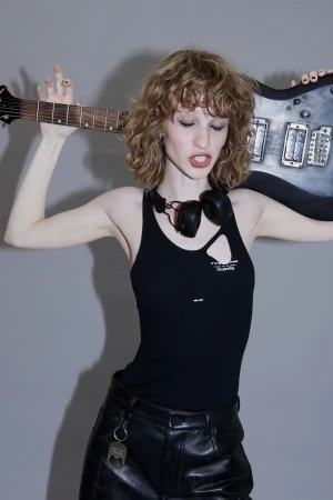Model with curly hair in a black tank top and leather pants holding an electric guitar against a grey seamless background