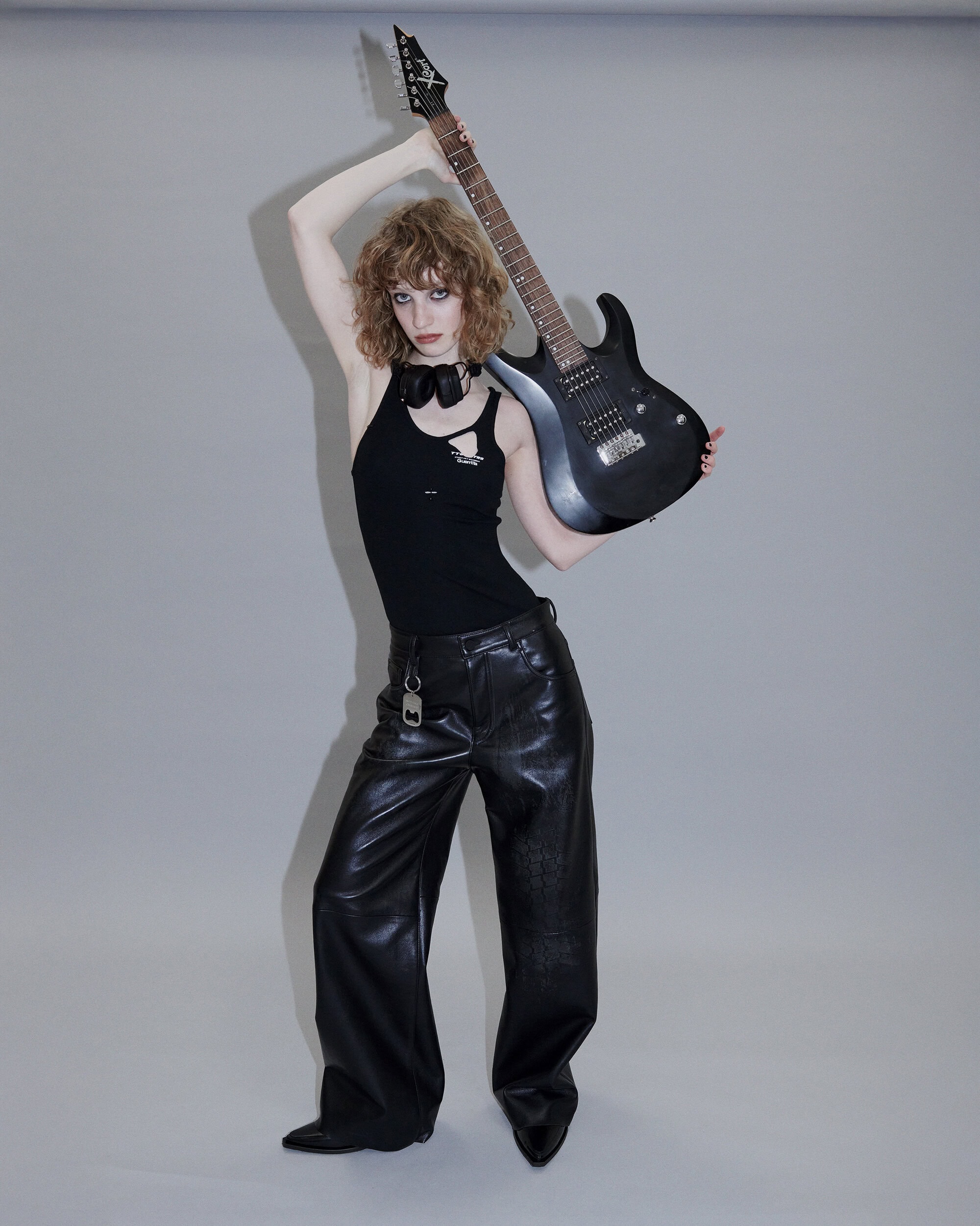 Model in black tank top and shiny leather trousers holding an electric guitar against a plain grey studio seamless background.