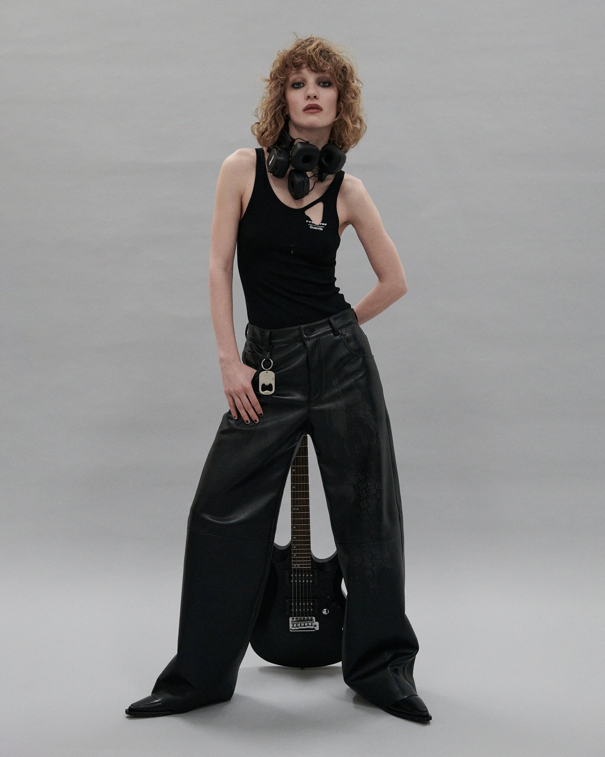 Model with curly red hair wearing a black tank top and black leather trousers standing against a seamless grey backdrop.