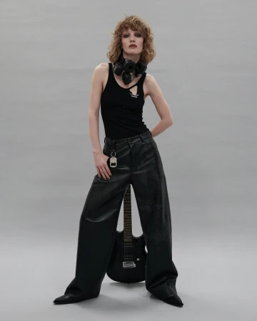 Model with curly red hair wearing a black tank top and black leather trousers standing against a seamless grey backdrop.