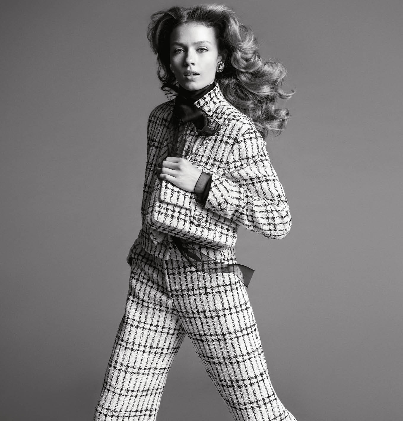 Model wearing a tweed suit and holding a quilted handbag against a seamless grey backdrop for the Chanel Spring 2025 ad campaign.