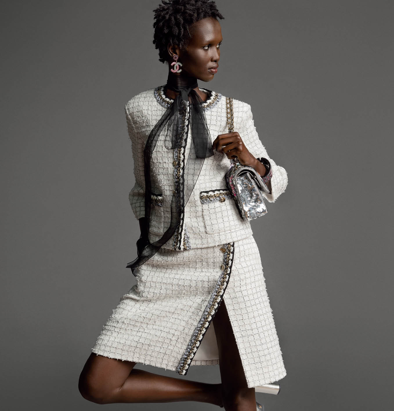 Model wearing a white tweed Chanel skirt suit with a black sheer bow tie and holding a sequined handbag