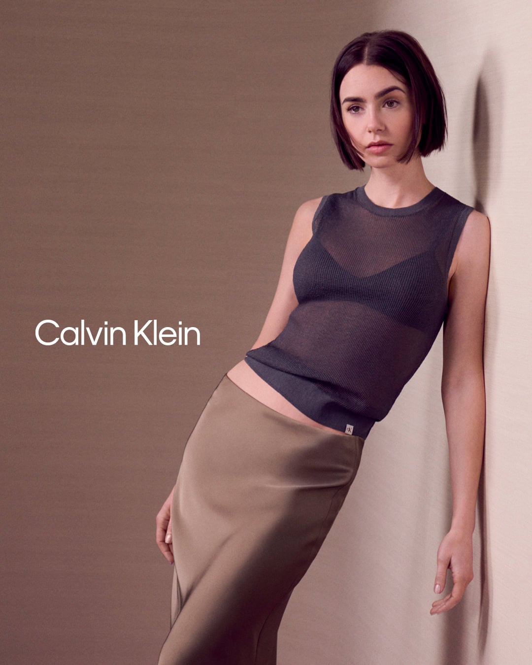 Lily Collins leaning against a neutral wall wearing a sheer dark grey sleeveless top and a long satin skirt for Calvin Klein.