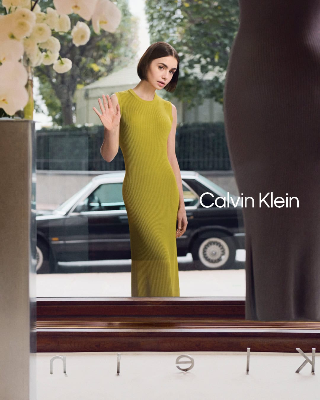 Lily Collins in a yellow sleeveless dress waving inside a storefront window for Calvin Klein spring 2025 ad campaign.