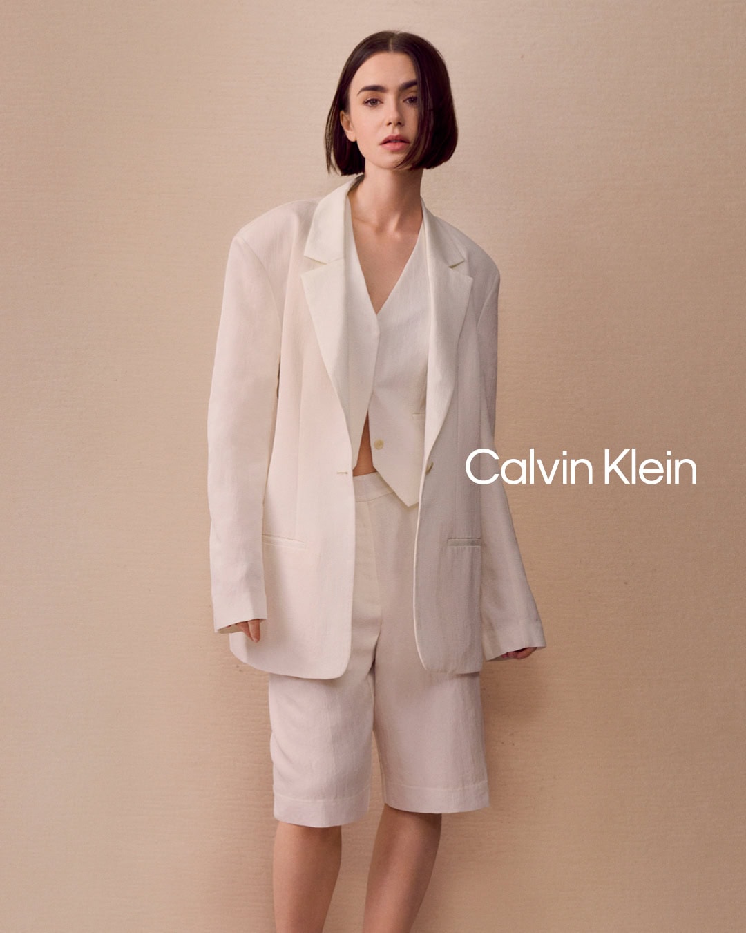 Lily Collins in a white blazer and matching shorts against a textured neutral background for Calvin Klein.