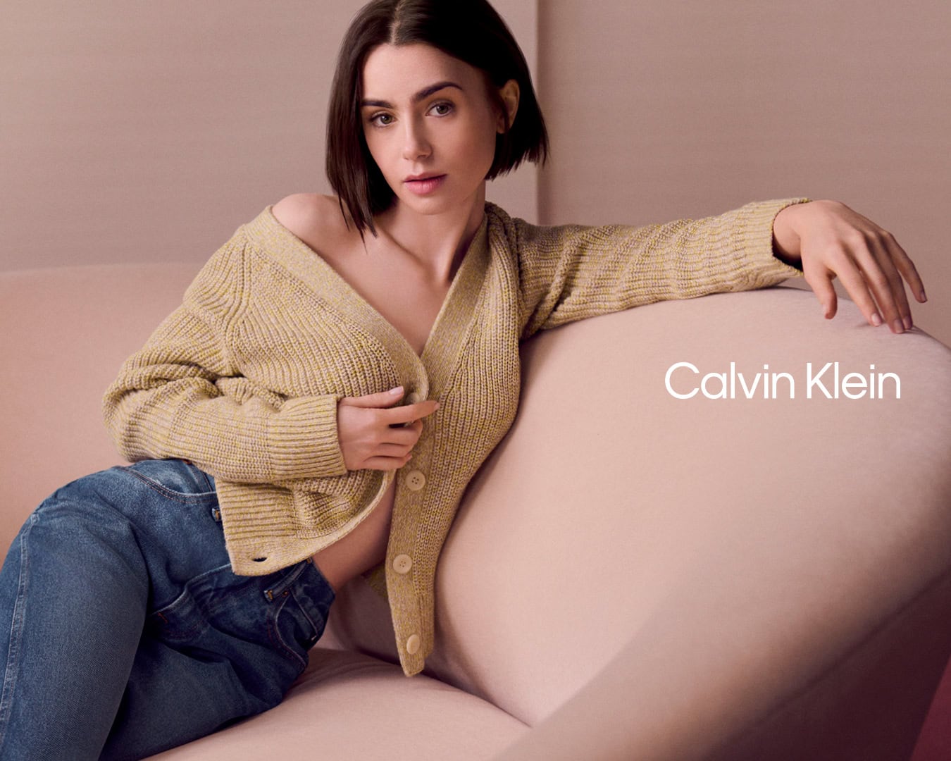 Lily Collins wearing a yellow cardigan and blue jeans, seated on a pink sofa in a Calvin Klein ad campaign.