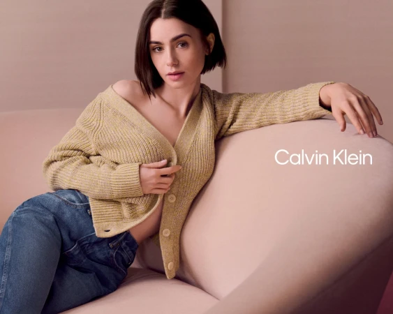 Lily Collins wearing a yellow cardigan and blue jeans, seated on a pink sofa in a Calvin Klein ad campaign.