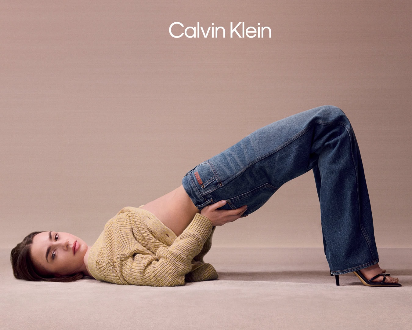 Model Lily Collins arching backwards in a beige knit cardigan and blue denim jeans against a seamless beige backdrop.