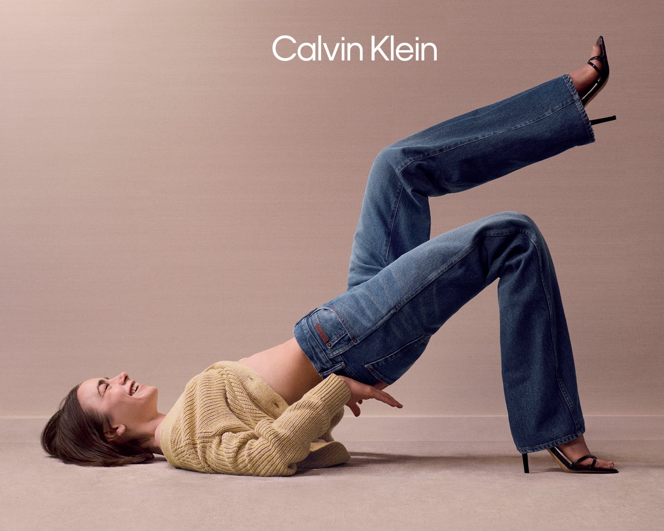 Lily Collins wearing a yellow cardigan and blue jeans posing playfully on a neutral seamless studio background for Calvin Klein.