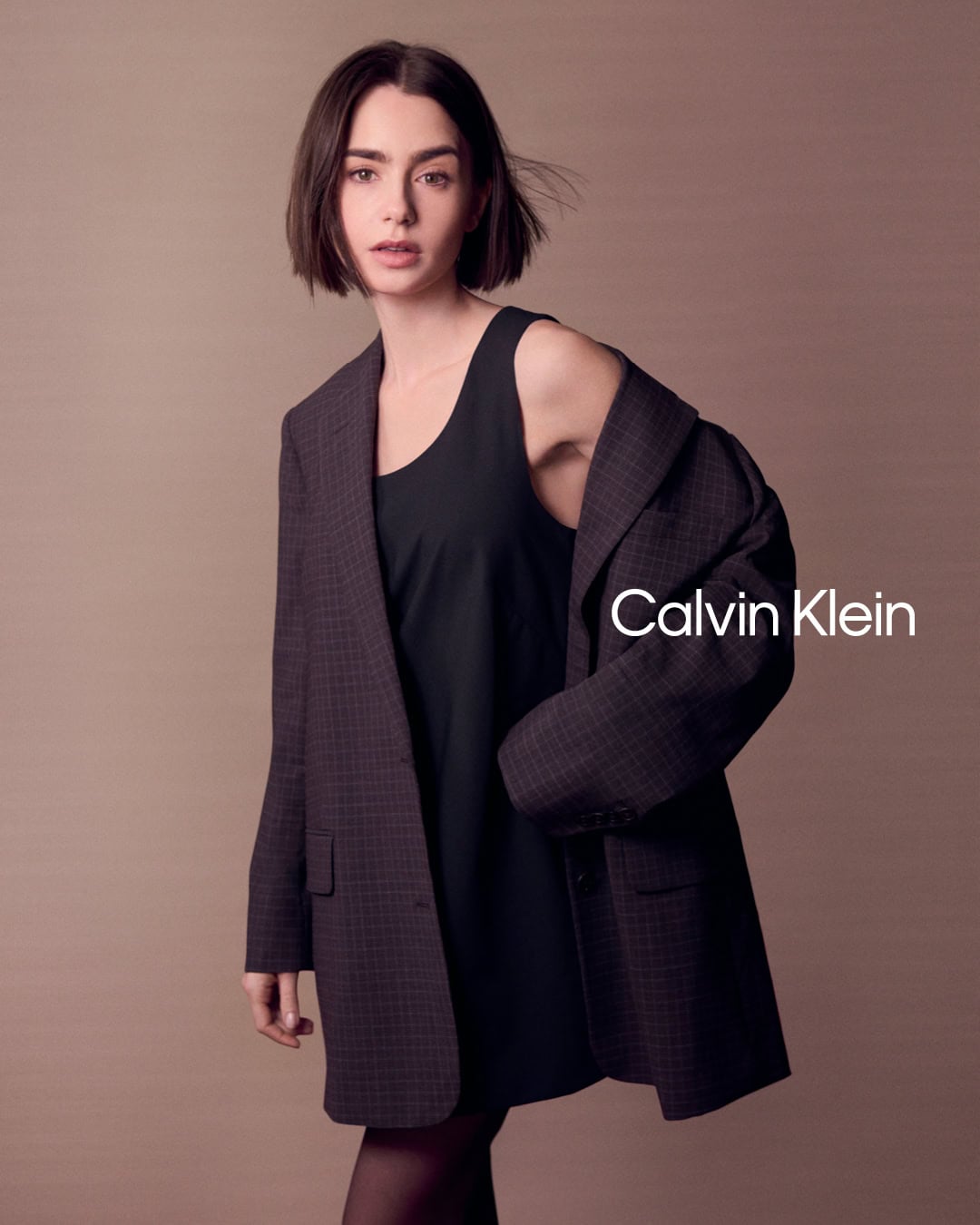 Lily Collins wearing a dark blazer over a black dress against a plain brown background for Calvin Klein