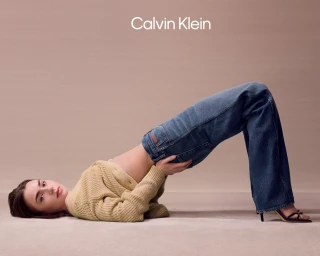 Model Lily Collins arching backwards in a beige knit cardigan and blue denim jeans against a seamless beige backdrop.