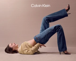 Lily Collins wearing a yellow cardigan and blue jeans posing playfully on a neutral seamless studio background for Calvin Klein.