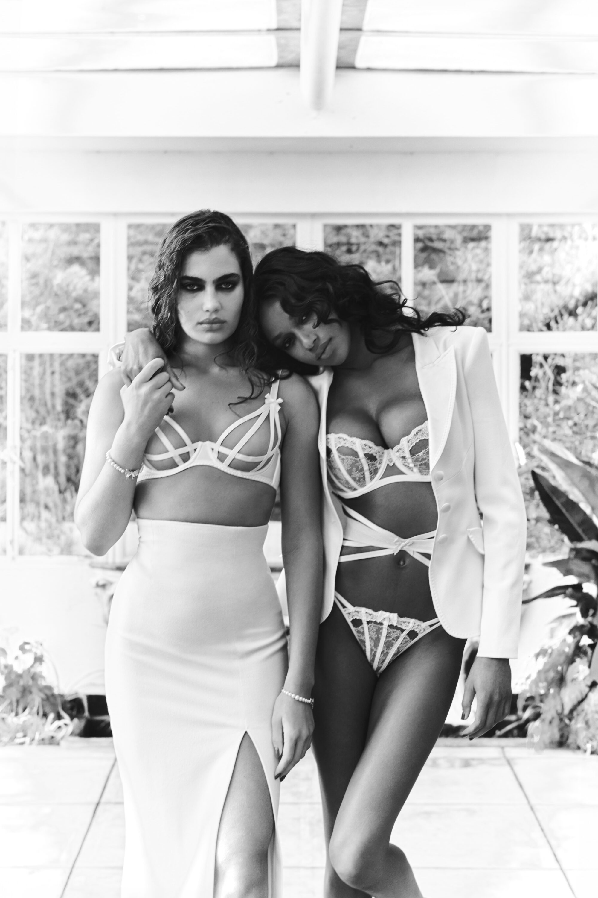 Black and white portrait of two women wearing white lace lingerie and a blazer standing indoors near a large window