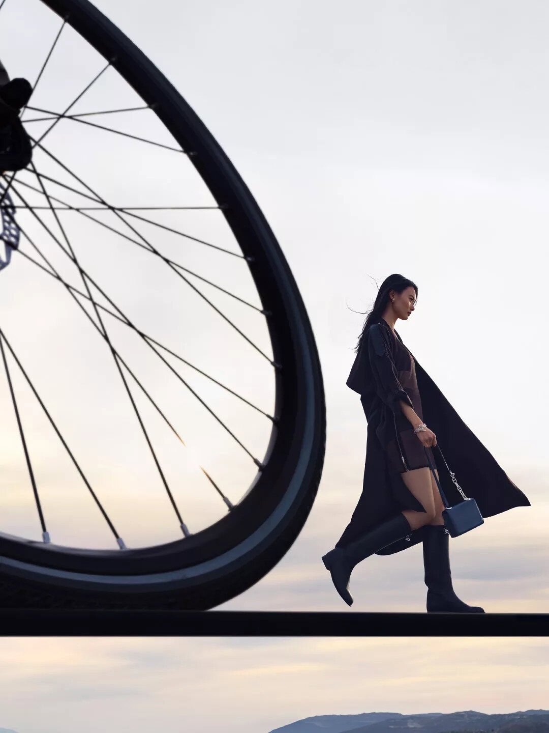 Model walking against an overcast sky with a giant bicycle wheel in the foreground for the Hermes Spring 2025 ad campaign.