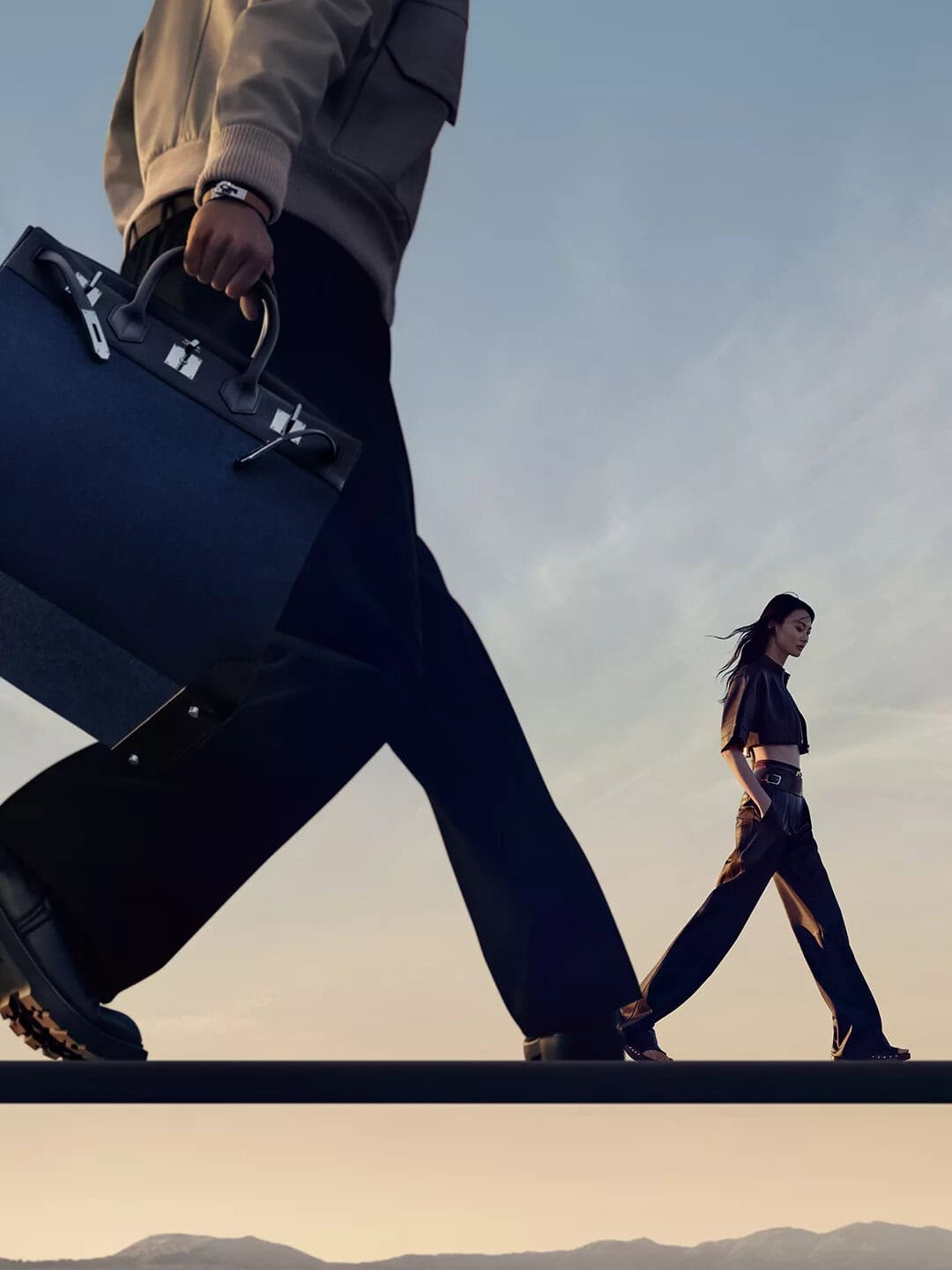 Low angle shot of a person carrying a large dark handbag with another model walking in the distance against a sunset sky