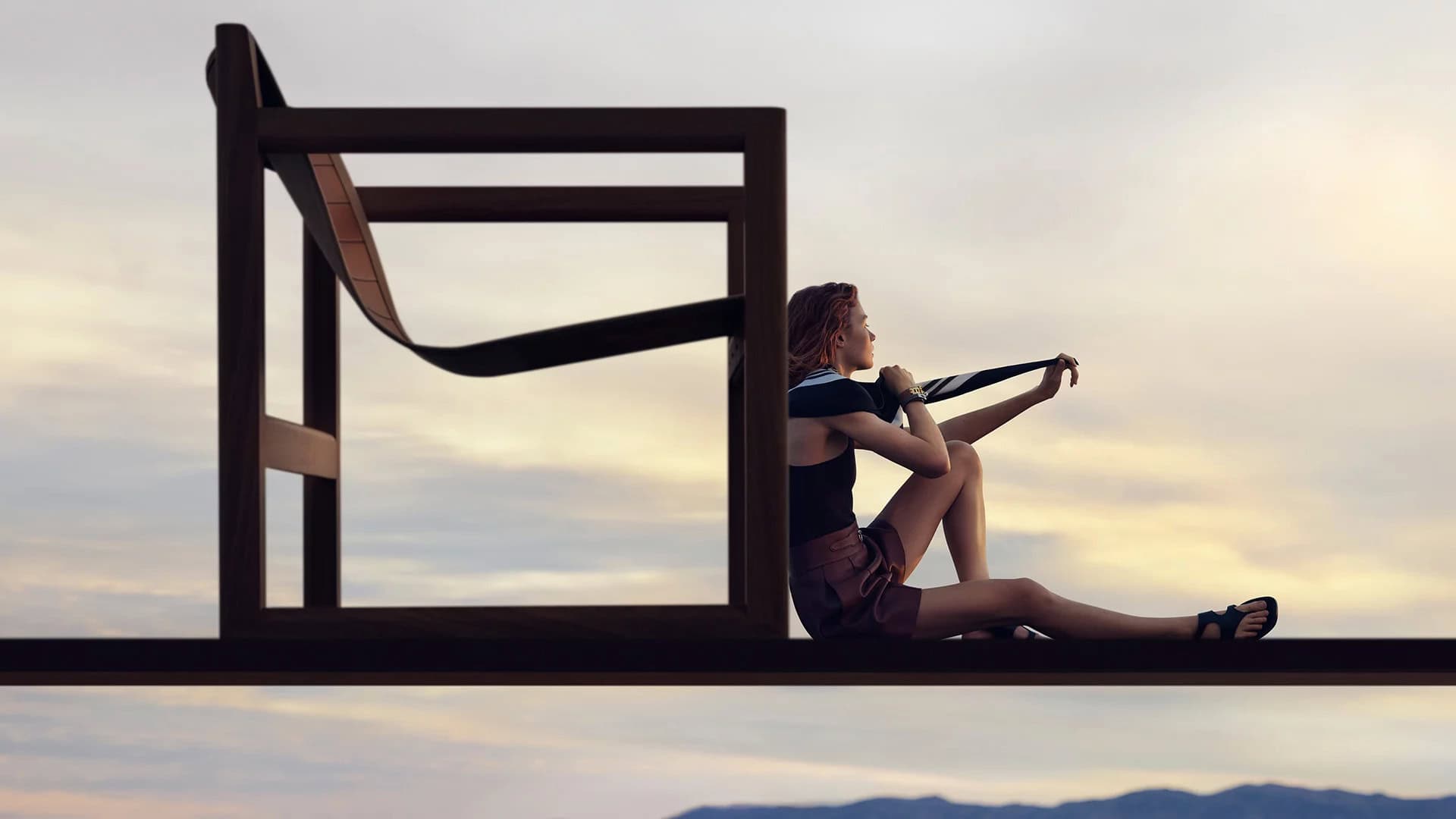 Woman seated on an elevated platform next to a large minimalist wooden chair frame against a pale sky