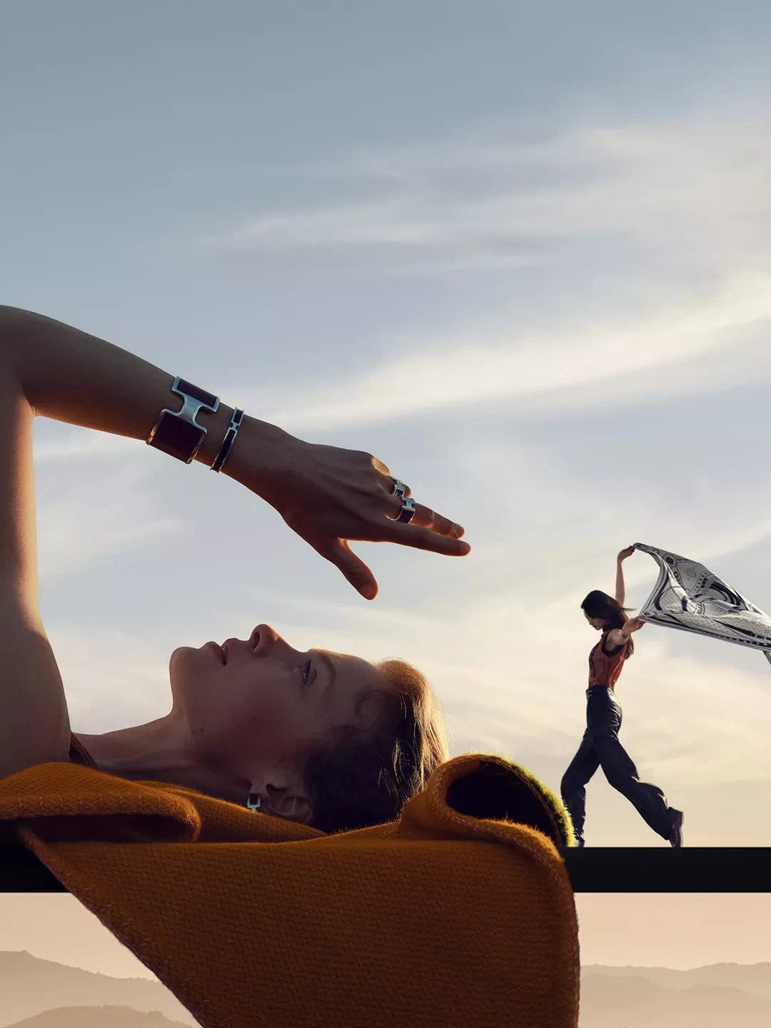 Close-up profile of a woman lying down with bracelets, next to a small figure holding a scarf against a sky background.