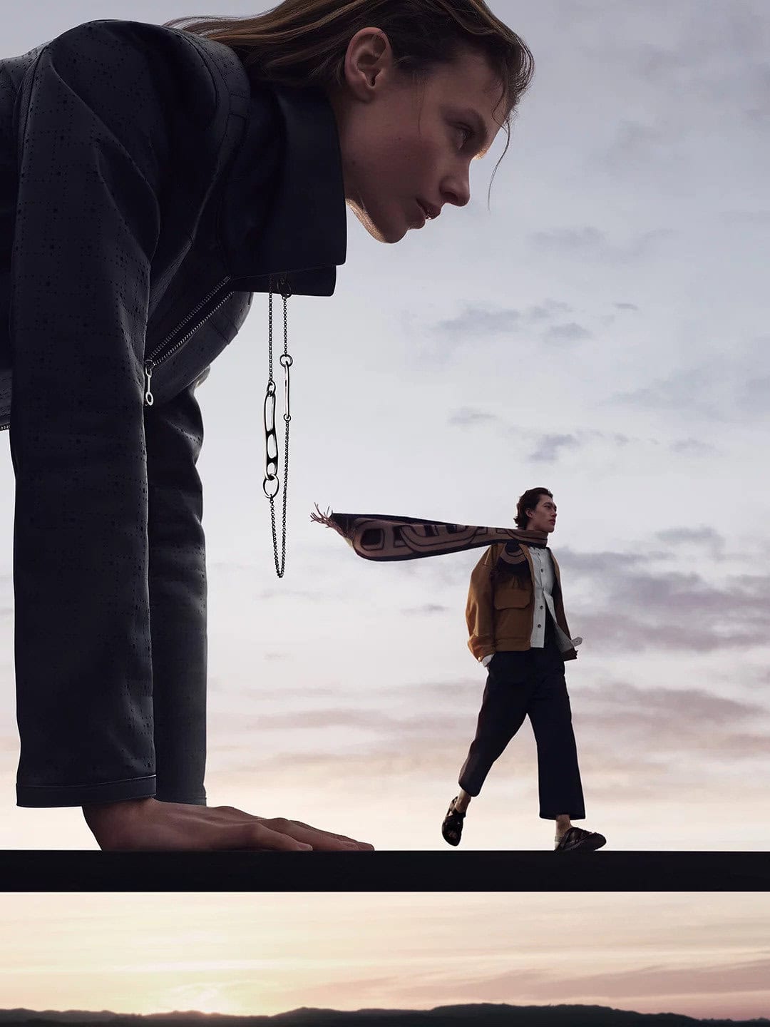 Hermes Spring 2025 ad campaign featuring a woman leaning forward with a smaller man walking in a landscape against an overcast sky.