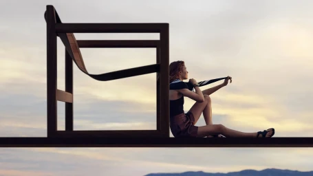 Woman seated on an elevated platform next to a large minimalist wooden chair frame against a pale sky