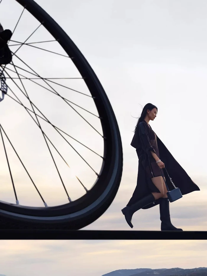 Model walking against an overcast sky with a giant bicycle wheel in the foreground for the Hermes Spring 2025 ad campaign.