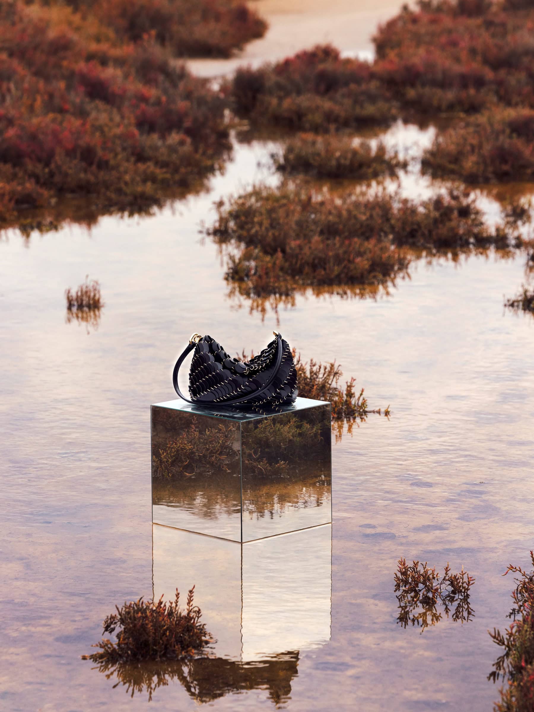 Black studded handbag resting on a mirrored cube pedestal set in shallow water surrounded by vegetation.