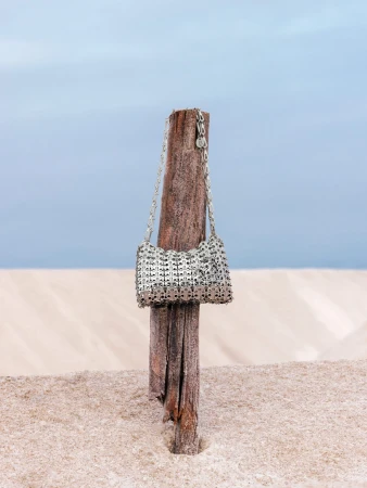 Silver chainmail handbag hanging on a weathered wooden post against a sandy beach and soft blue sky background.
