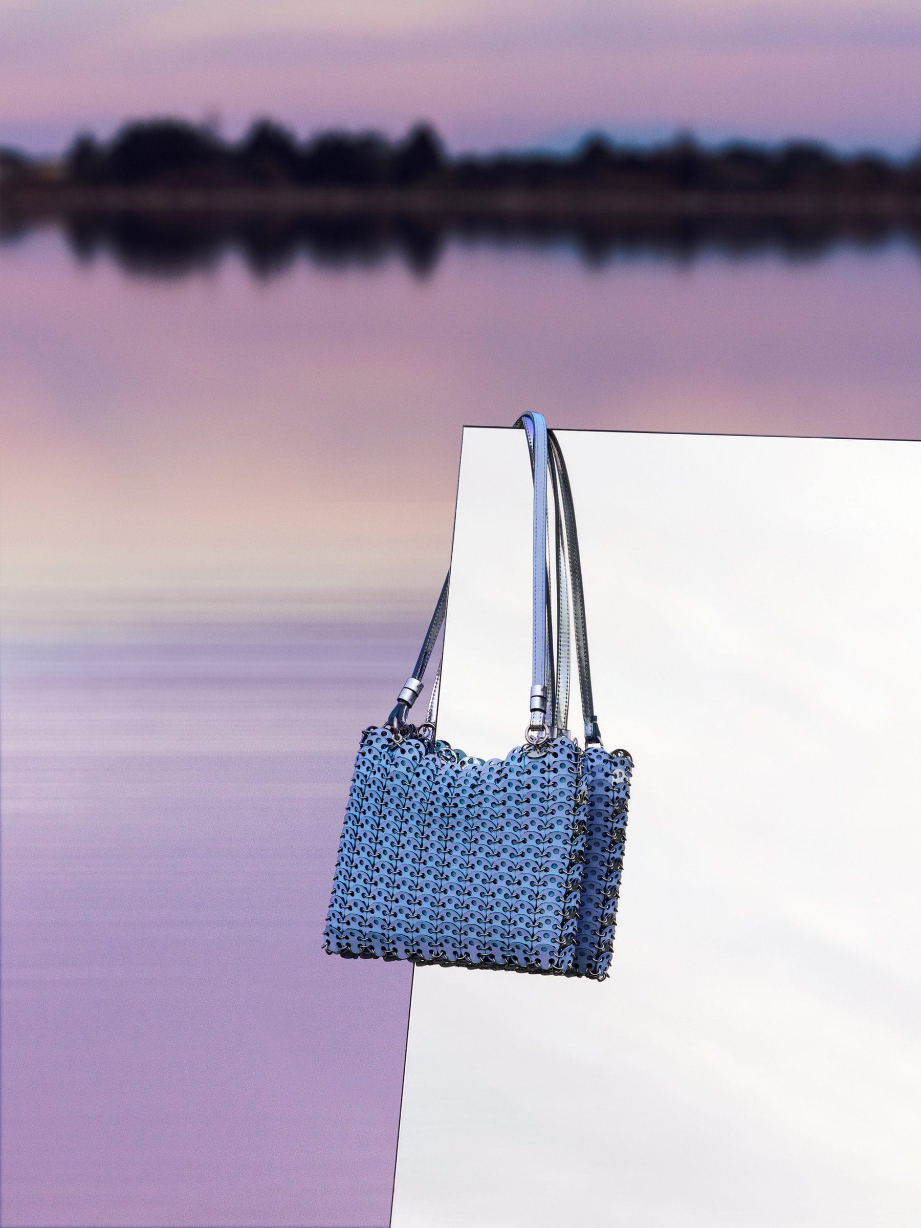 Blue metallic chainmail handbag hanging against a white panel with a sunset water backdrop for Rabanne