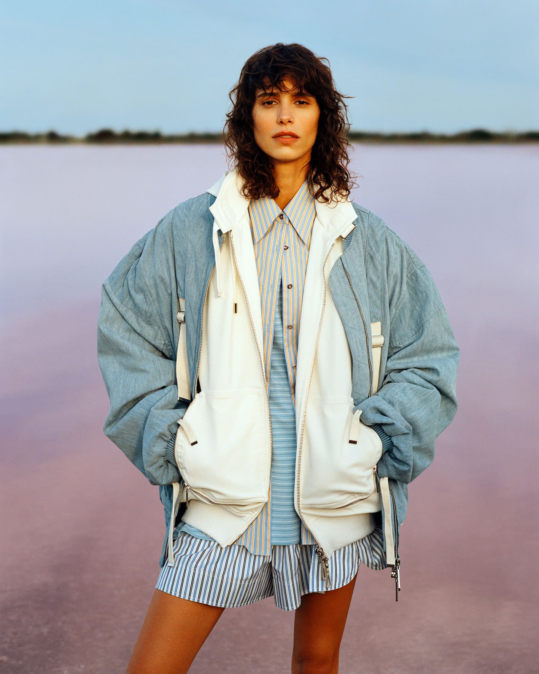 Model standing outdoors wearing a blue jacket, white layered vest, and striped shirt against a pink water horizon.