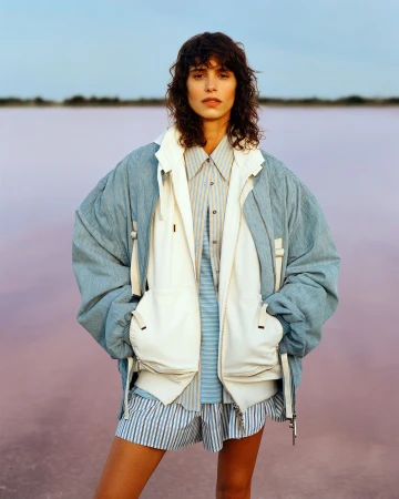 Model standing outdoors wearing a blue jacket, white layered vest, and striped shirt against a pink water horizon.