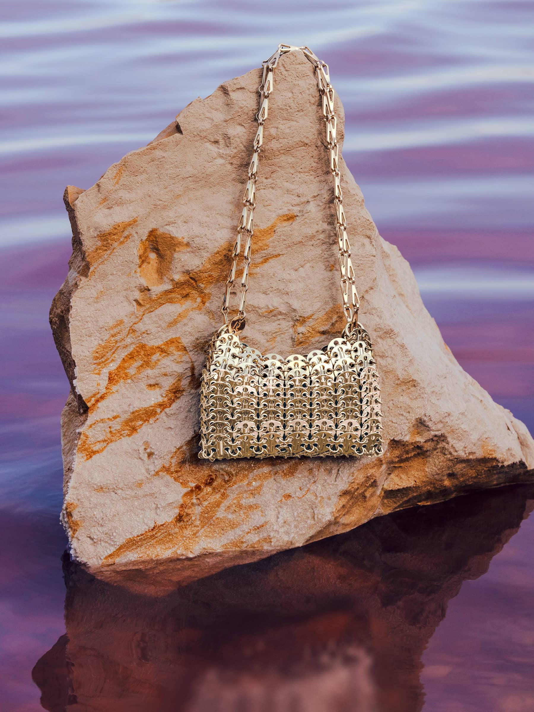 Gold metallic chainmail handbag resting on a jagged light brown rock above calm purple water