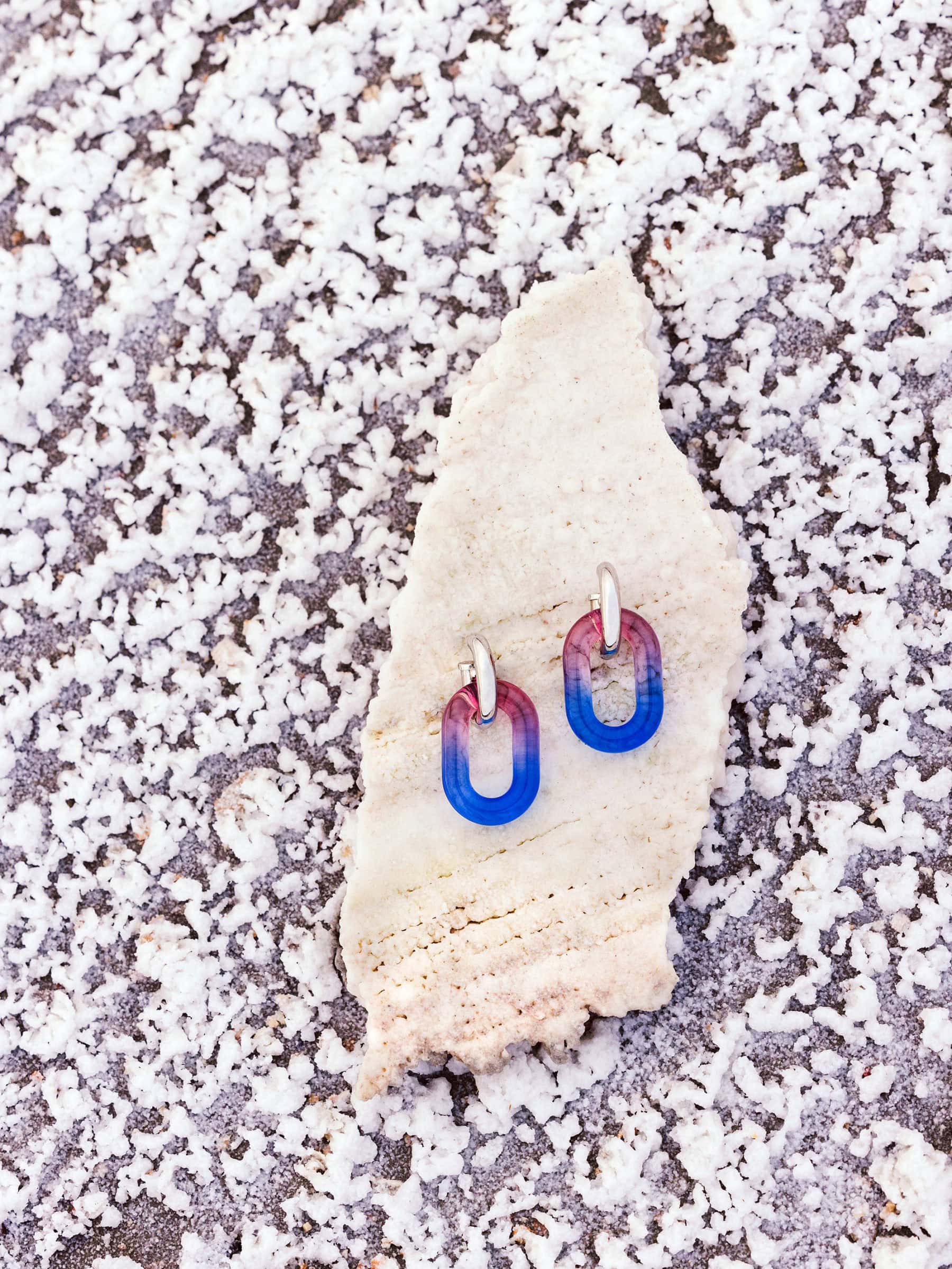 Close-up product shot of Rabanne gradient blue and pink chain link earrings resting on a pale textured stone surface.