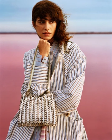 Model Mica Arganaraz wearing a striped coat holding a metallic mesh handbag outdoors against a pink water background