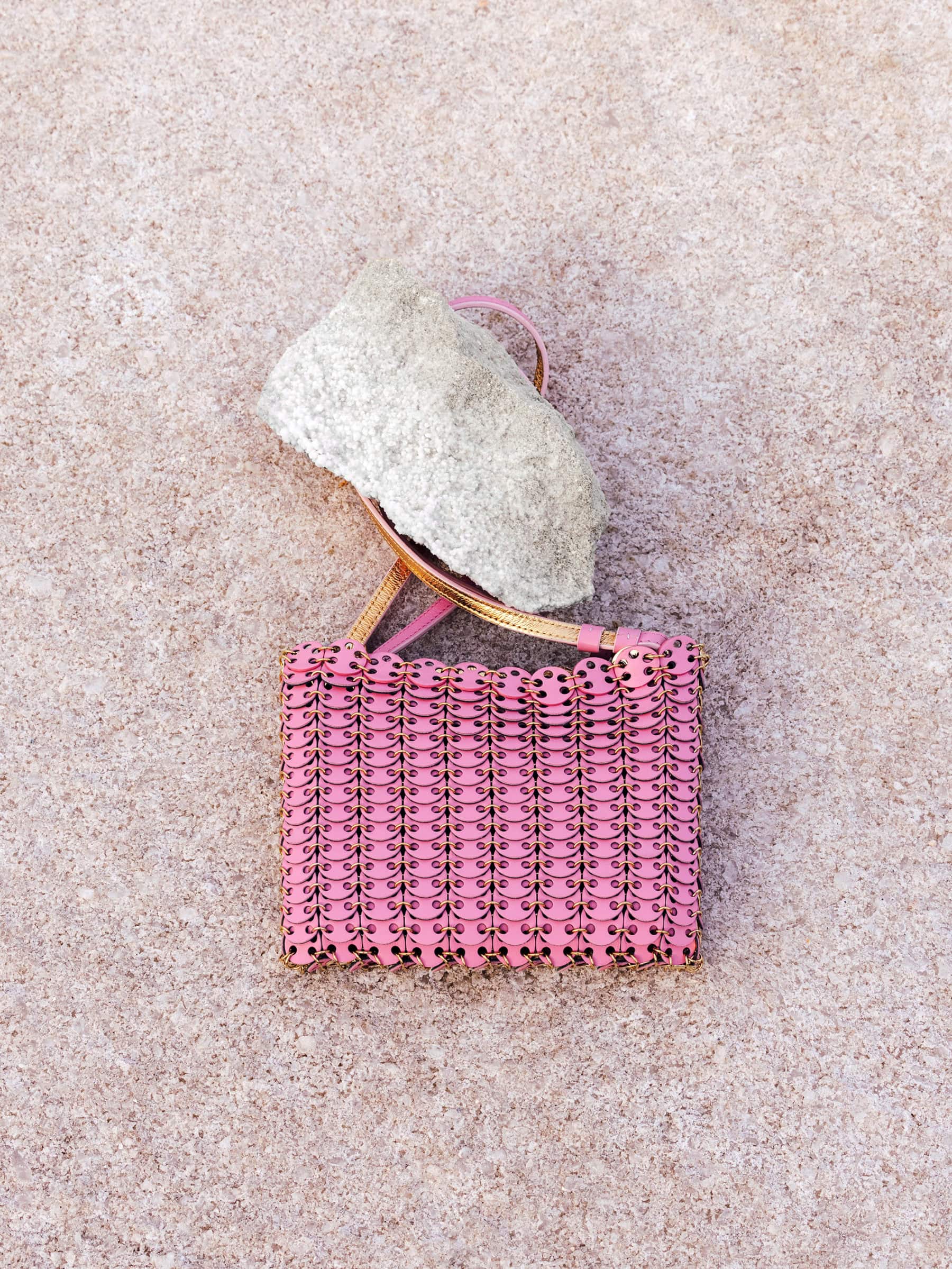 Pink chainmail handbag with a thin strap resting on a textured concrete surface alongside a rough gray stone.